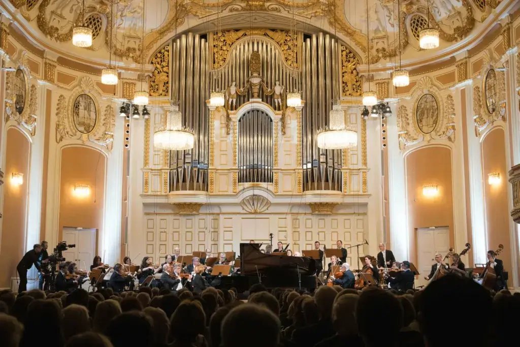 The image captures a moment from the opening concert of Mozart Week 2025, featuring a performance by the Mozarteum Orchestra Salzburg, conducted by Roberto González-Monjas. It includes renowned artists such as pianists Martha Argerich and Rolando Villazón. The photo, taken by Wolfgang Lienbacher, showcases the musicians on stage, with the orchestra in full performance, creating a captivating and dynamic atmosphere.