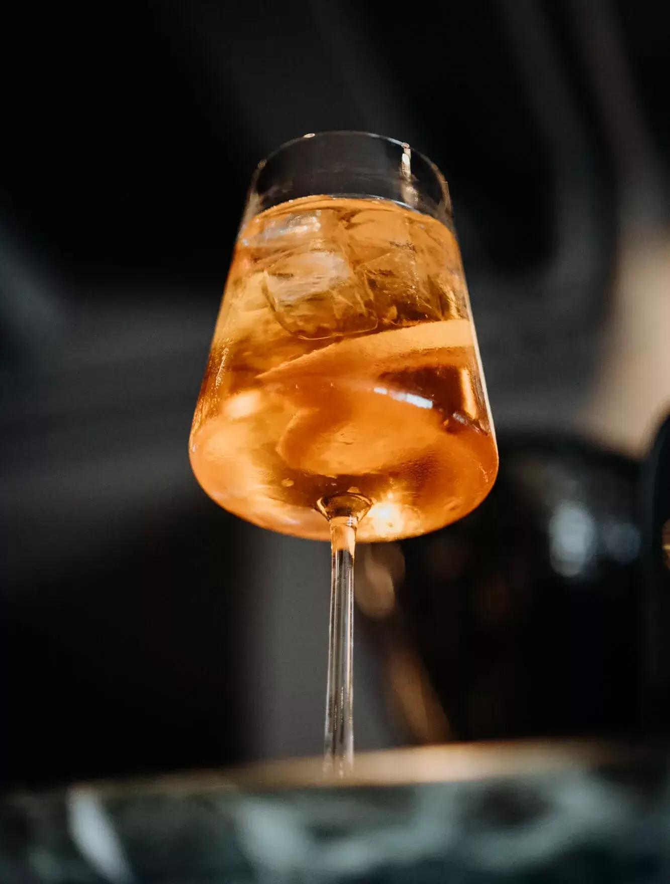 A view from below of a glass of Aperol Spritz on the bar, showcasing the vibrant orange color of the drink, with bubbles rising and the glass reflecting the ambient lighting around it.