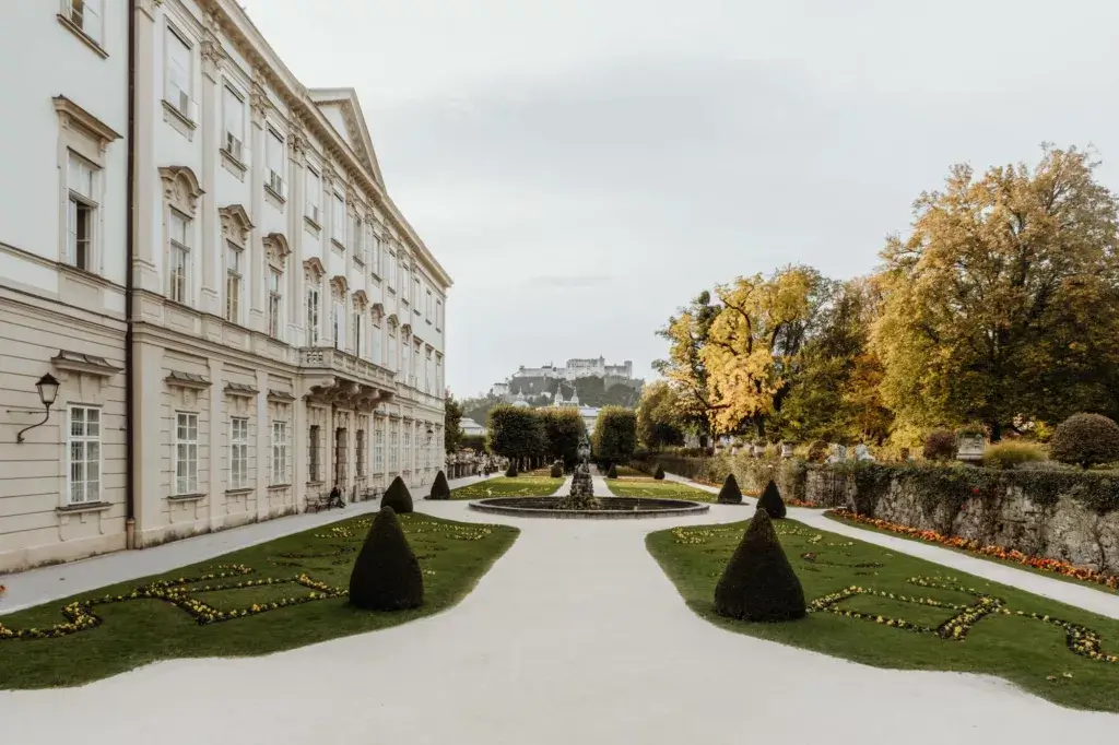 A view of the Mirabell Garden in Salzburg, with its meticulously arranged flower beds, fountains, and sculptures, offering a stunning backdrop of the Hohensalzburg Fortress. The lush greenery and vibrant flowers create a peaceful atmosphere, while the fortress towers above the city in the distance.