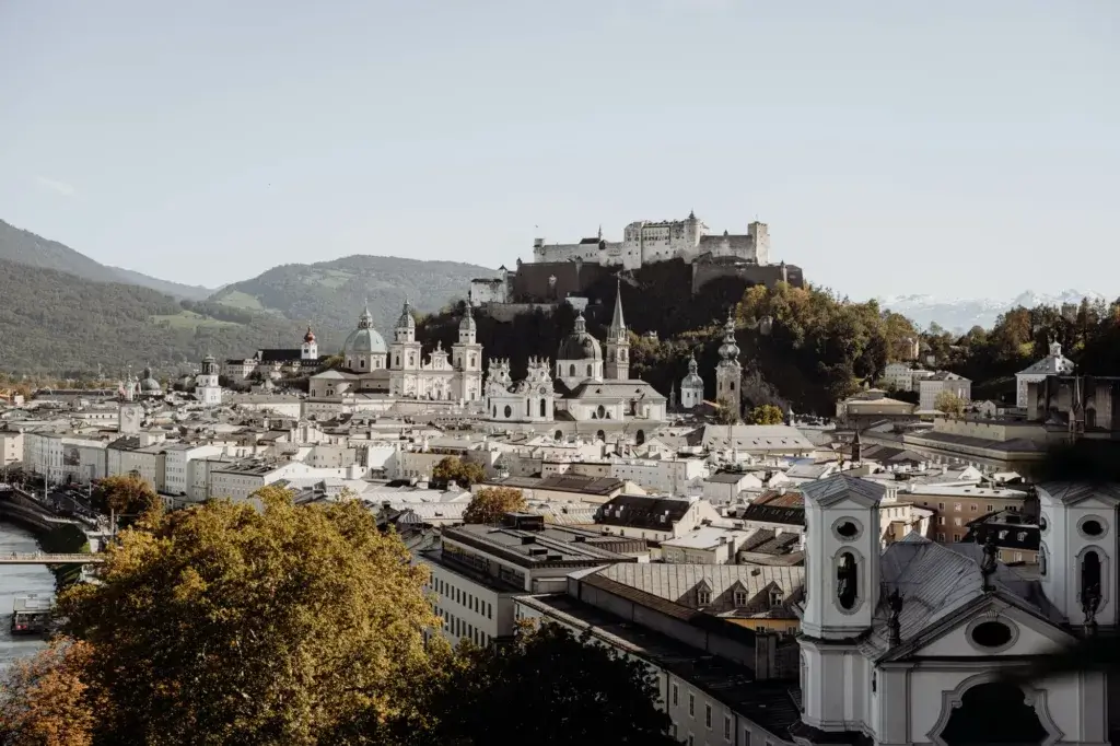 A panoramic view over the city of Salzburg, with its charming rooftops and historic buildings, leading up to the iconic Hohensalzburg Fortress perched atop the hill. The backdrop features the stunning Alpine mountains, creating a beautiful contrast with the city's classical architecture.