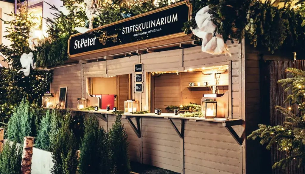 A Christmas-themed hut at the St. Peter Christmas Market in Salzburg, adorned with festive decorations and lights. The cozy wooden stall offers holiday treats, creating a warm and inviting atmosphere amid the winter charm of Salzburg's Christmas market.